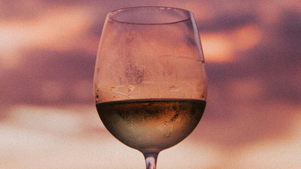 Primo piano di un calice di vino trasparente stagliato contro uno suggestivo cielo estivo sfumato di rosa e viola al crepuscolo, l'atmosfera delle Notti del vino.